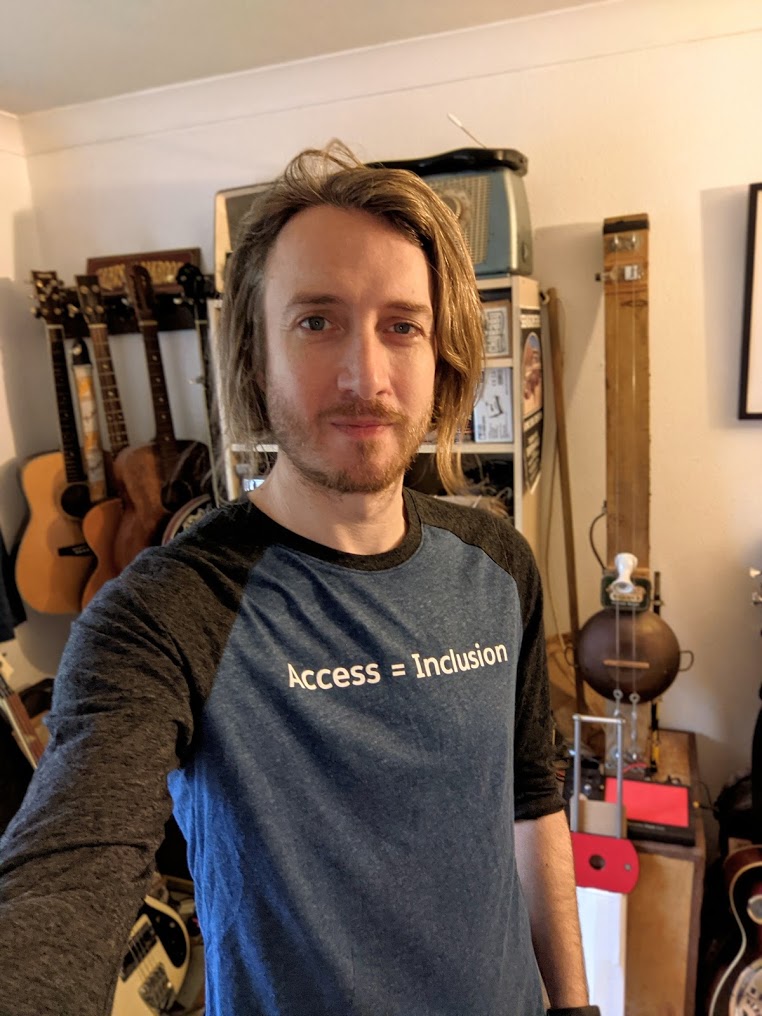 Mark in a blue t-shirt that read an access = inclustion. in the backgroudn there are several guitars including a DIY 2 sting diddley bow.